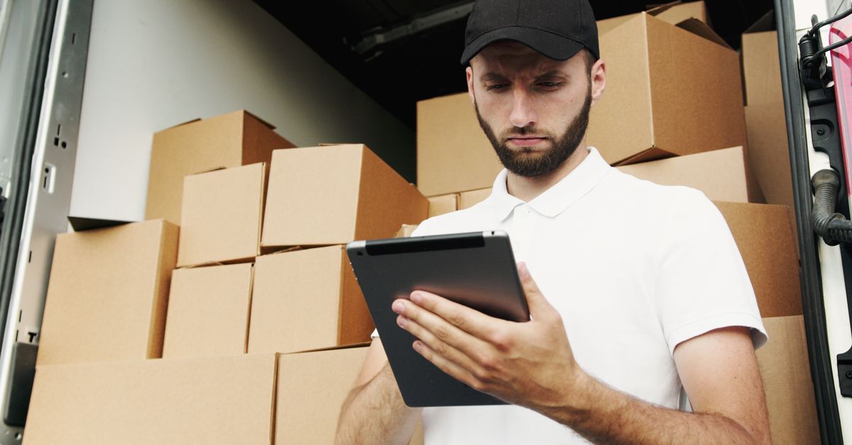 Delivery worker using a tablet to manage shipments with stacked boxes in the background.